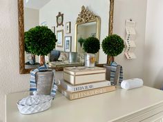 a white dresser topped with books and vases filled with green topiaries next to a mirror