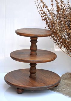 three tiered wooden cake stand next to a vase with dried flowers in it on a white surface