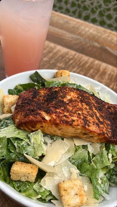 a white plate topped with salad and meat next to a pink drink on top of a wooden table