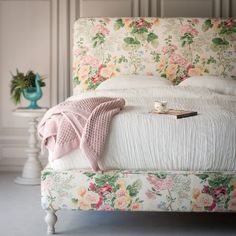 a white bed topped with a pink blanket next to a blue vase filled with flowers