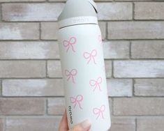 a hand holding a white and pink water bottle with bows on it, against a brick wall