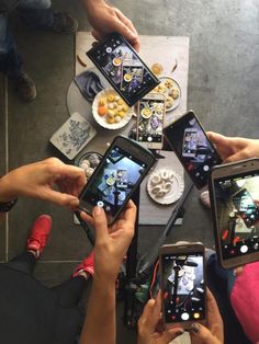 four people holding up their cell phones in the middle of a table with food on it