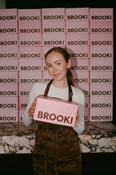 a woman holding up a box with the word brook on it in front of a wall of pink boxes