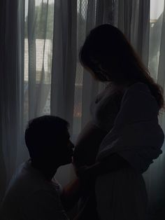 a pregnant woman standing next to a man in front of a window with sheer curtains