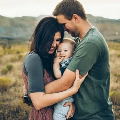 a man and woman holding a baby in the middle of a field with mountains in the background