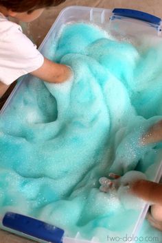 two children are playing with blue foam in a plastic container on the floor and one child is reaching for something