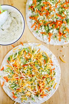 two pizzas with cheese, broccoli and carrots next to a bowl of dip