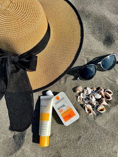 a hat, sunglasses and sunscreen sitting on the sand next to some seashells