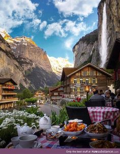 a table with food on it in front of some mountains and buildings at the base of a waterfall