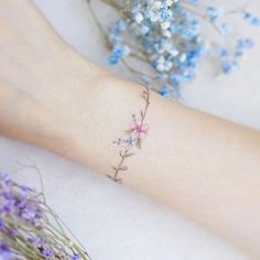 a small flower tattoo on the left wrist and right arm, with blue flowers in the background