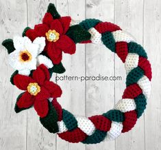 a crocheted christmas wreath with flowers and leaves on the front, sitting on a white wooden surface