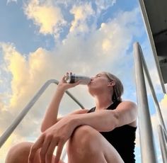 a woman drinking from a water bottle while sitting on the side of a metal railing