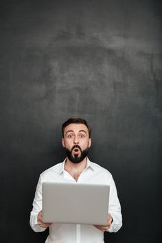 a man with a beard holding a laptop in front of his face and looking surprised