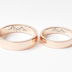 two rose gold wedding bands with mountains engraved on the sides, set against a white background