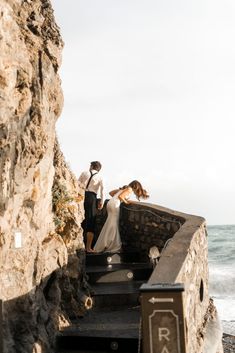 a man and woman standing on top of a stone wall next to the ocean