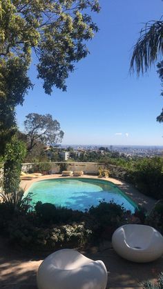 an outdoor swimming pool surrounded by greenery and trees, with two bean shaped lounge chairs in the foreground