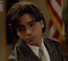 a young man wearing glasses and a leather jacket looks at the camera while sitting in front of an american flag