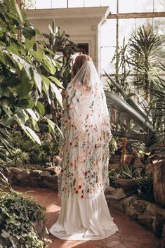 a woman in a white wedding dress and veil walking through a greenhouse filled with plants