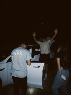 two people standing next to each other in front of a white car with their hands up