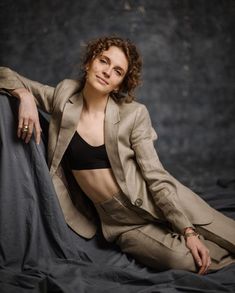 a woman sitting on top of a bed wearing a tan suit and black bralet