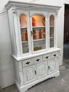 an old white china cabinet with glass doors and cupboards on the bottom, in front of a wall