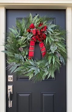 a christmas wreath hanging on the front door with evergreen leaves and red plaid bow tied around it