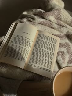 an open book sitting on top of a blanket next to a cup of coffee