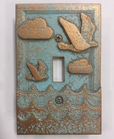 a decorative light switch cover with birds flying over the water and clouds in gold on blue