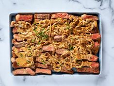 a pan filled with meat and noodles on top of a marble countertop next to a knife