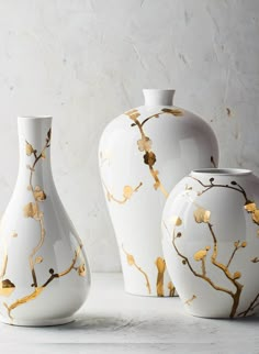 three white vases with gold leaf designs on them sitting next to each other in front of a marble wall