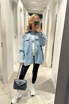 A stylish maternity look featuring a woman wearing an oversized light-wash denim jacket over a white top, paired with black leggings. She accessorizes with white chunky sneakers, white crew socks, and a black quilted handbag with a chain strap. Her blonde hair is styled in loose waves, and she holds her phone in front of her face, taking a mirror selfie in a well-lit fitting room with white walls and tiled floors. Cozy Outfits Fall, Looks With Leggings, Pregnancy Winter Outfits, Cute Fall Outfit Ideas, Fall Fashion Outfit Ideas, Pregnancy Fashion Winter, Pregnancy Fashion Fall