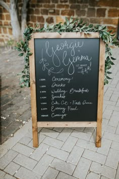 a chalkboard sign with greenery on it sitting in front of a brick wall