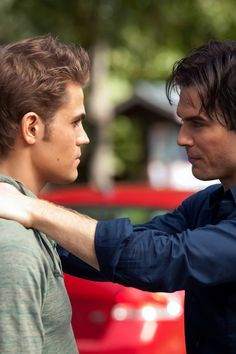 two young men standing next to each other in front of a red car and one is adjusting the collar of another man's shirt