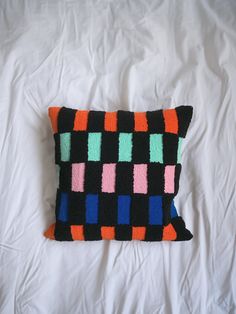 a black pillow with multicolored squares on it sitting on a white bed sheet