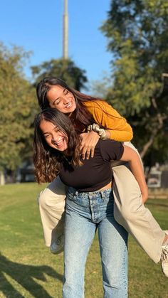 two women are smiling and hugging in the park with their arms around each other as they stand on one another's shoulders