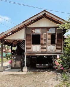 an old wooden house sitting on the side of a dirt road next to trees and flowers