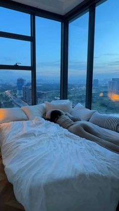 a person laying on top of a white bed in a bedroom next to large windows