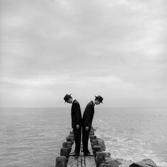 two men in suits and hats standing on a pier over the ocean with their backs to each other