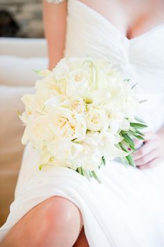 a bridal holding a bouquet of white flowers and greenery in her hand, sitting on a couch