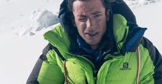 a man in a green jacket standing on top of a snow covered slope with his mouth open