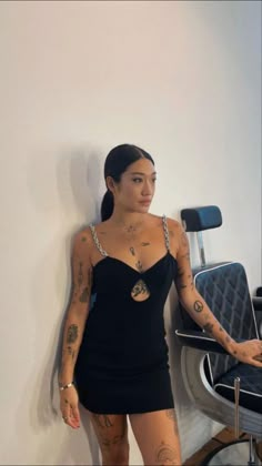 a woman standing next to a chair with tattoos on her arms and legs, wearing a black dress