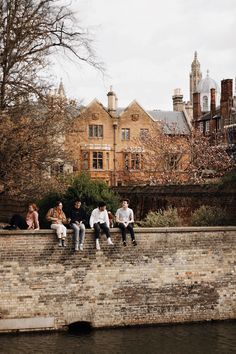 four people sitting on a brick wall next to a body of water with buildings in the background