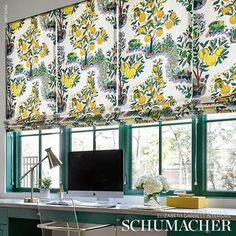 a desk with a computer on top of it next to two windows covered in roman shades