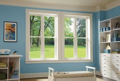 a bedroom with blue walls and white furniture in the foreground is a large open window that looks out onto a grassy field