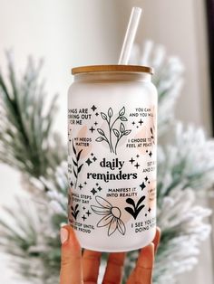 a person holding up a white cup with the words daily reminders on it and a pine tree in the background