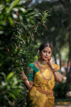 a woman in a yellow and green sari standing next to some trees with her hands on her chin