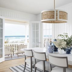 a dining room table with white chairs and vases on top of it in front of the ocean