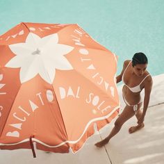 a woman standing next to a swimming pool holding an orange and white umbrella over her head