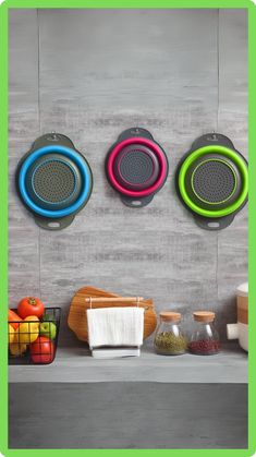 three colorful strainers are hanging on the wall next to some fruit and other items