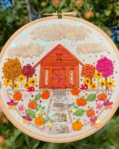 a hand holding up an embroidered hoop with a house and flowers in the center on it